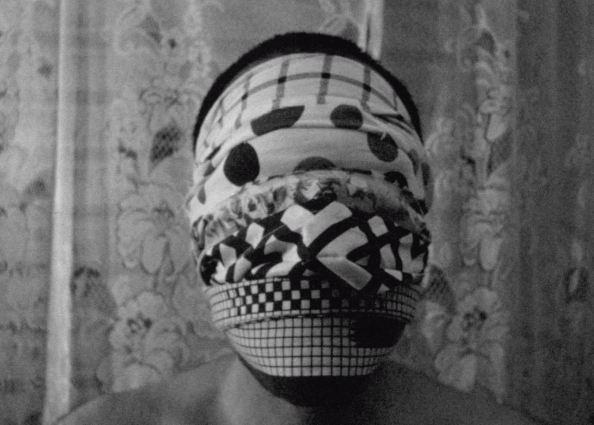 Black and white, analog, 16mm photo of a man’s face covered in patterned cloths. It appears as if he is wearing a mask. There is a floral curtain behind him. Shot with Bolex camera.