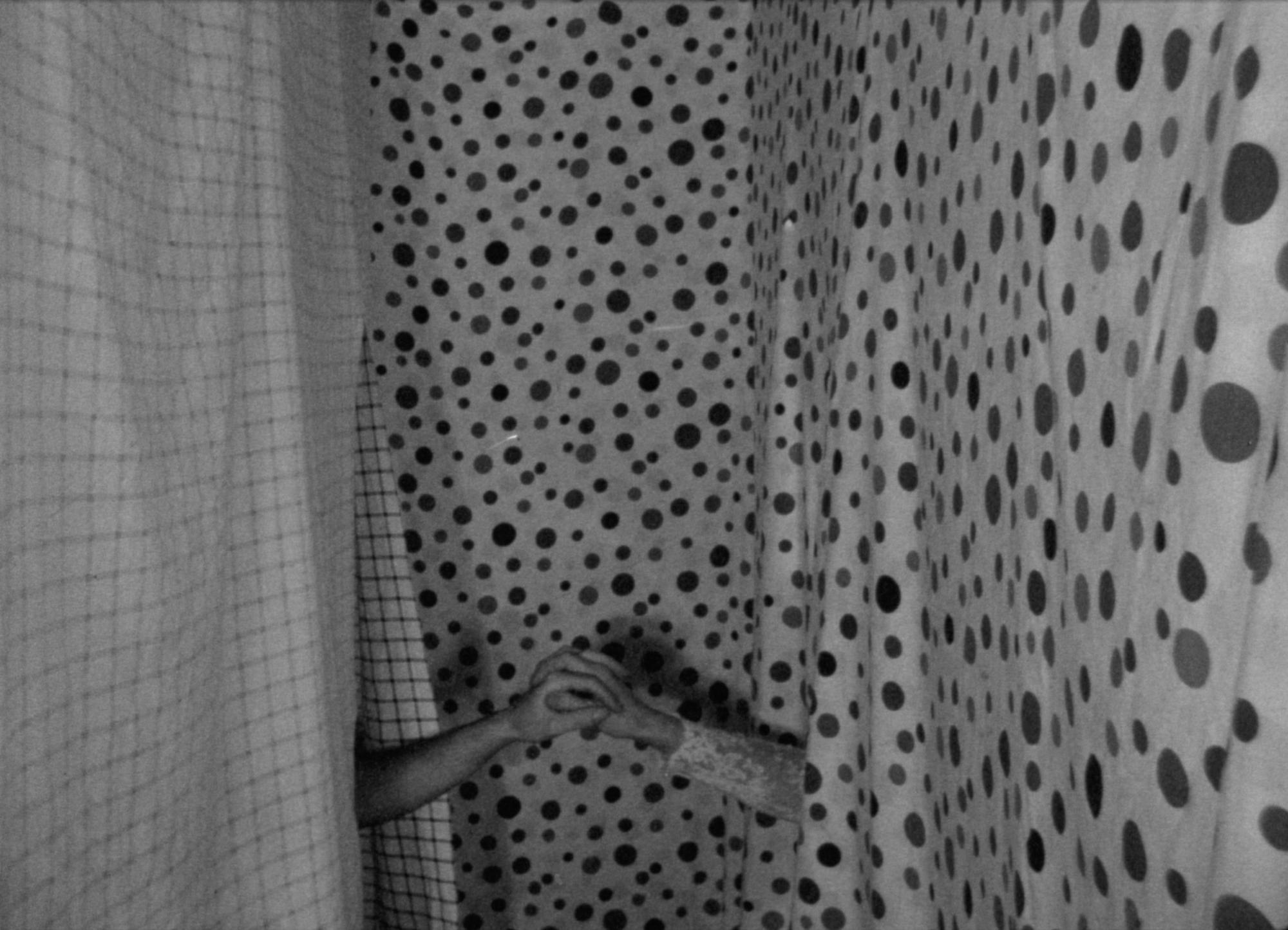 Black and white, analog, 16mm film still; two arms reach towards each other to hold hands in the center. They emerge from patterned curtains on the left and right side of the frame; one is checkered and the other is polka-dotted. Bolex footage.