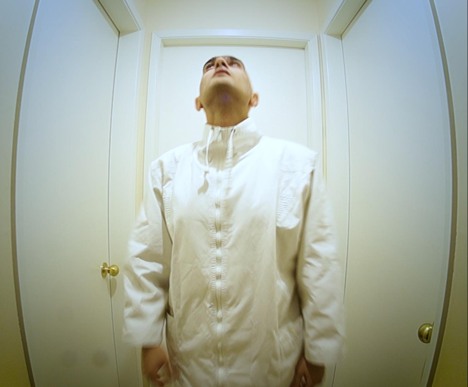 Man wearing all white stands looking upwards in the center of a small, surreal-like room with one door on each wall. 