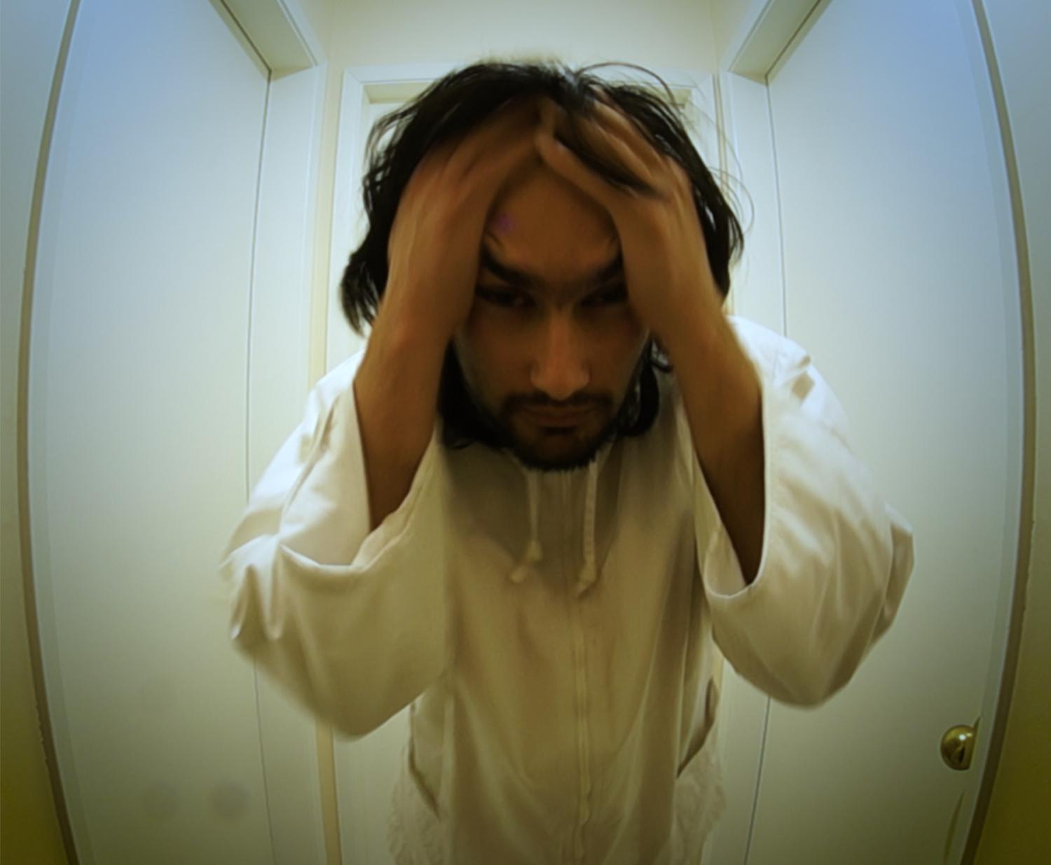 A bearded man wearing all white stands in the center of a small, dream-like room with one door on each wall. He faces the camera with both hands on his forehead; he has a worried expression.