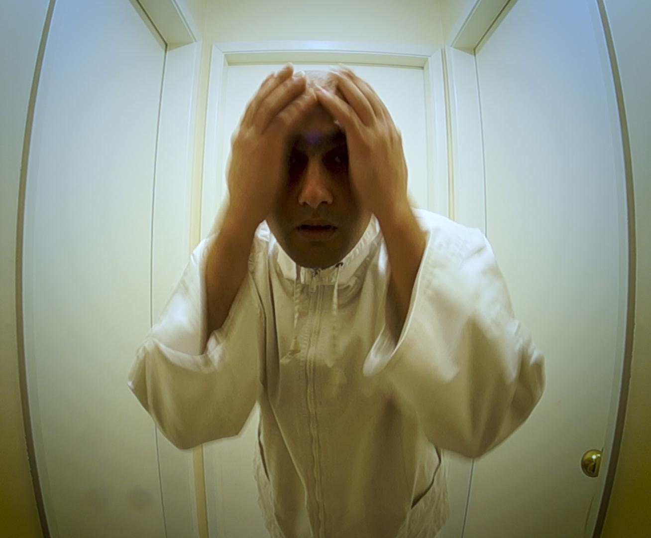 A bald man wearing all white stands in the center of a small, surreal room with one door on each wall. He faces the camera with both hands placed on his head; he has a fearful expression.