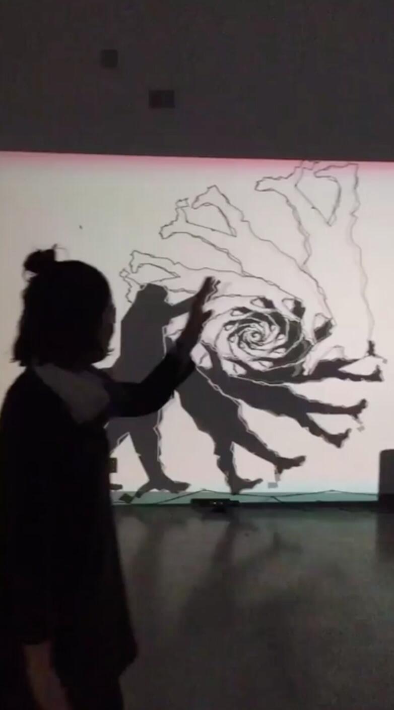 A girl interacts with an interactive video projection on a wall in front of her. There is a black and white vortex projecting on the wall, displaying an abstract replication of her silhouette.